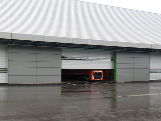 EFAFLEX high speed doors with bands of vision panels close a row of numbered loading bays on a wet forecourt, one door part raised with an orange lorry inside
