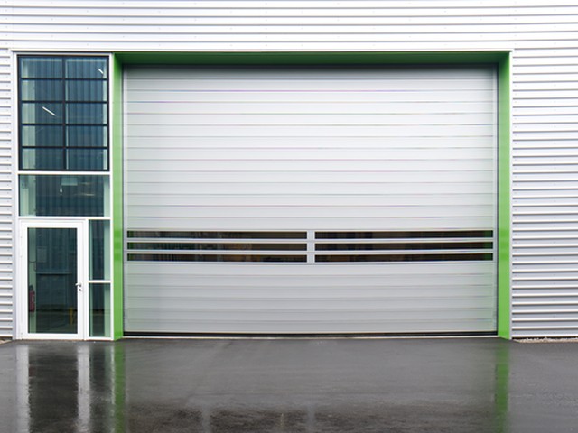 A large white EFAFLEX high speed door closed in the green framed opening of a metal clad unit, with a glazed personnel door alongside