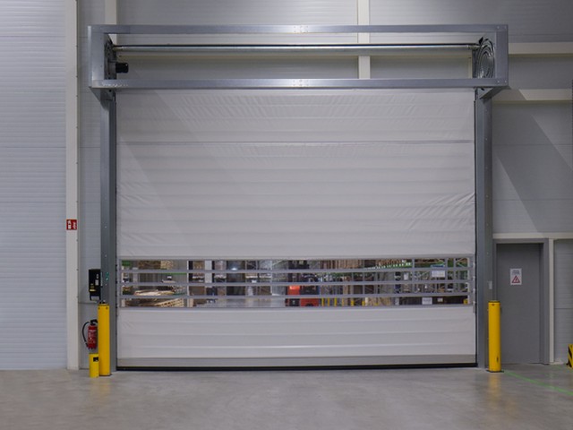 A tall white EFAFLEX high speed door closed in a hall, with a band of clear glazed slats across the middle and yellow bollards either side of the opening