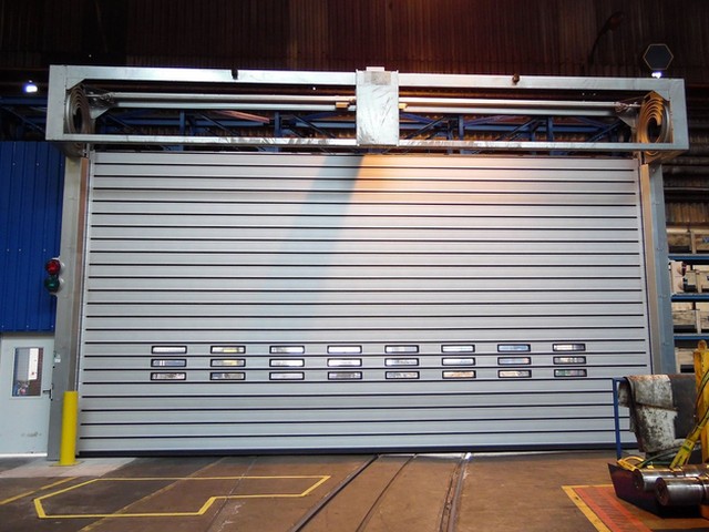Rails in the floor pass beneath a wide EFAFLEX high speed spiral door closed across a hall opening, vision windows across its leaf and the coil in the open headbox