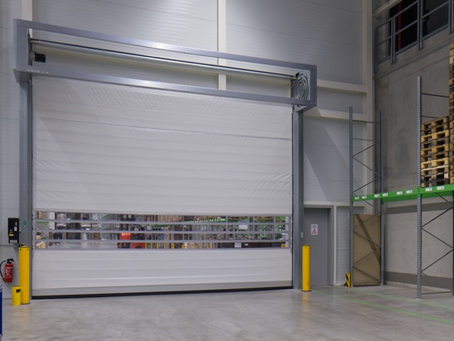 Pallet racking stands to one side of a large white EFAFLEX high speed door closed across an opening in a clean industrial hall, a glazed vision panel across its leaf