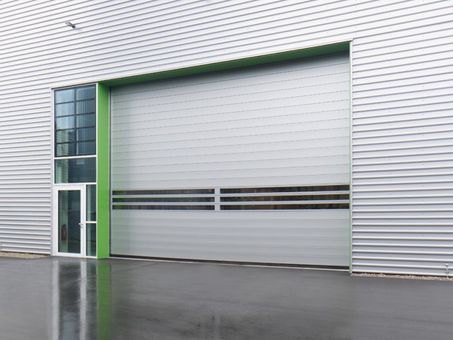 Set into the side of a profiled metal clad building, a large EFAFLEX high speed door sits closed in a green painted reveal beside a glazed personnel door