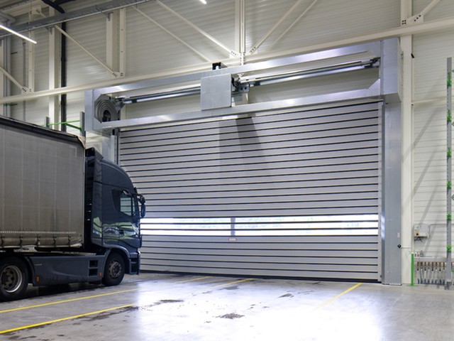 Inside an industrial hall, a large EFAFLEX high speed spiral door stands closed with a dark curtain sided lorry parked alongside