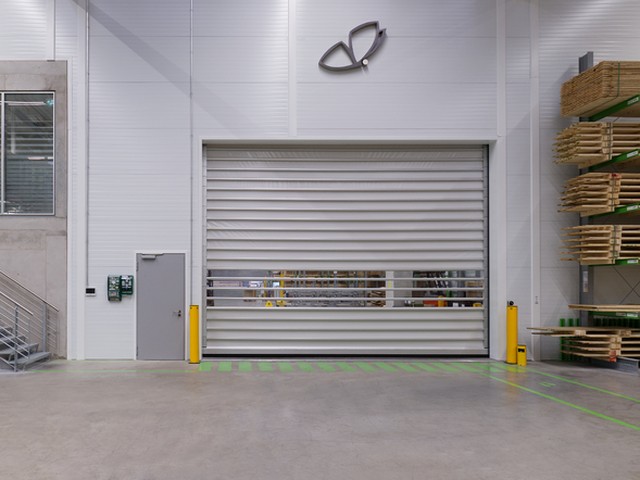 Closed inside a warehouse hall, a large EFAFLEX high speed door carries a band of vision panels across its leaf, with pallets stacked in racking alongside