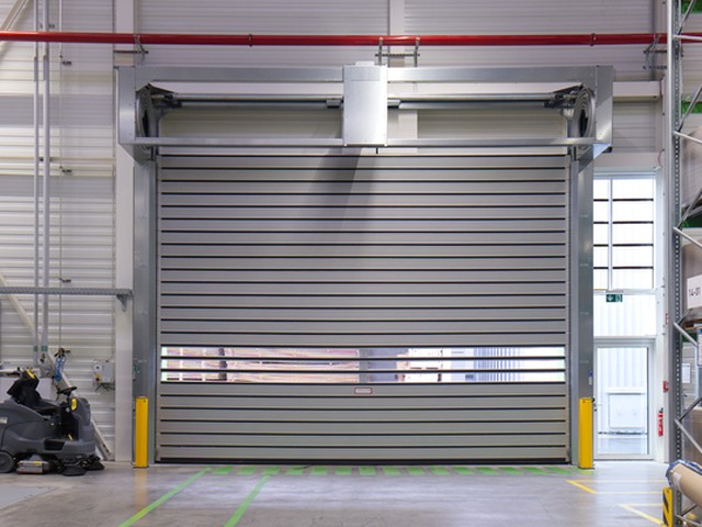 A large EFAFLEX high speed door closed inside a warehouse hall, a band of vision panels running across the leaf and yellow bollards either side