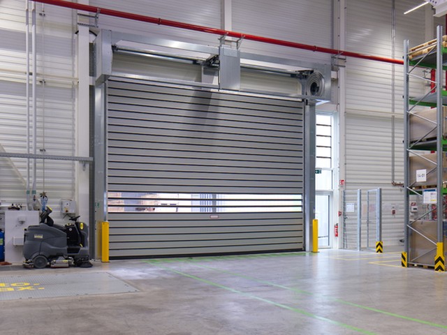 Yellow bollards flank the opening where a large EFAFLEX high speed spiral door sits closed in a warehouse hall, with loaded pallet racking to the right