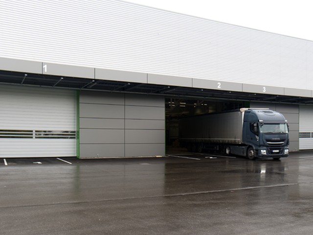 On a wet apron, a large EFAFLEX high speed door with a band of vision panels stands closed beside numbered bays, a dark lorry pulling out of the open one