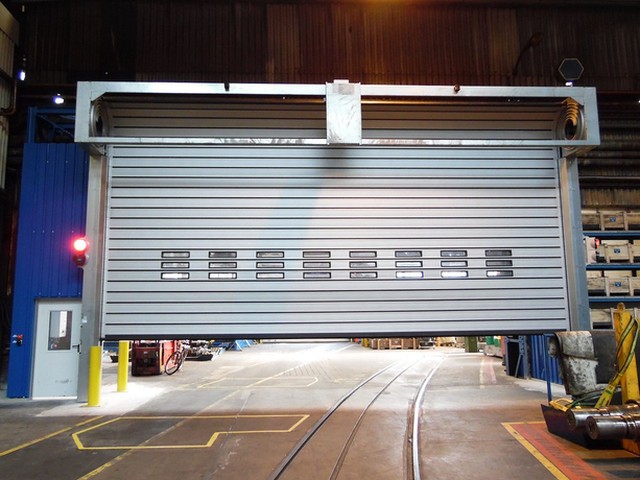 Raised above rail tracks in a workshop hall, a large EFAFLEX high speed door shows three rows of small vision windows across its leaf
